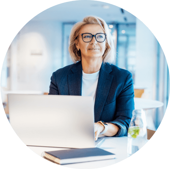 Actor portrayal of a woman with blond hair and glasses wearing a blue blazer sitting in front of an open laptop