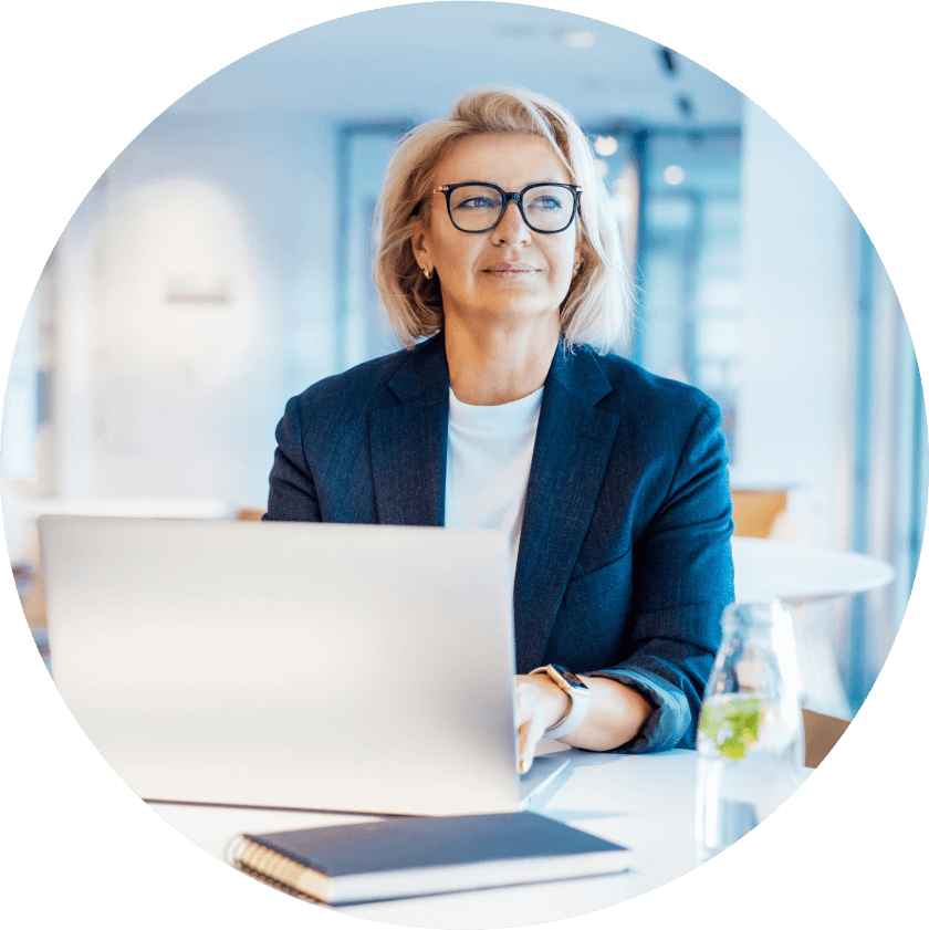 Actor portrayal of a woman with blond hair and glasses wearing a blue blazer sitting in front of an open laptop